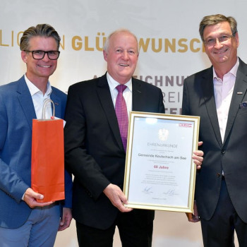 Ehrenurkunde für Gemeinde Keutschach am See wird von drei Männern in Anzug und Sakko entgegengenommen.